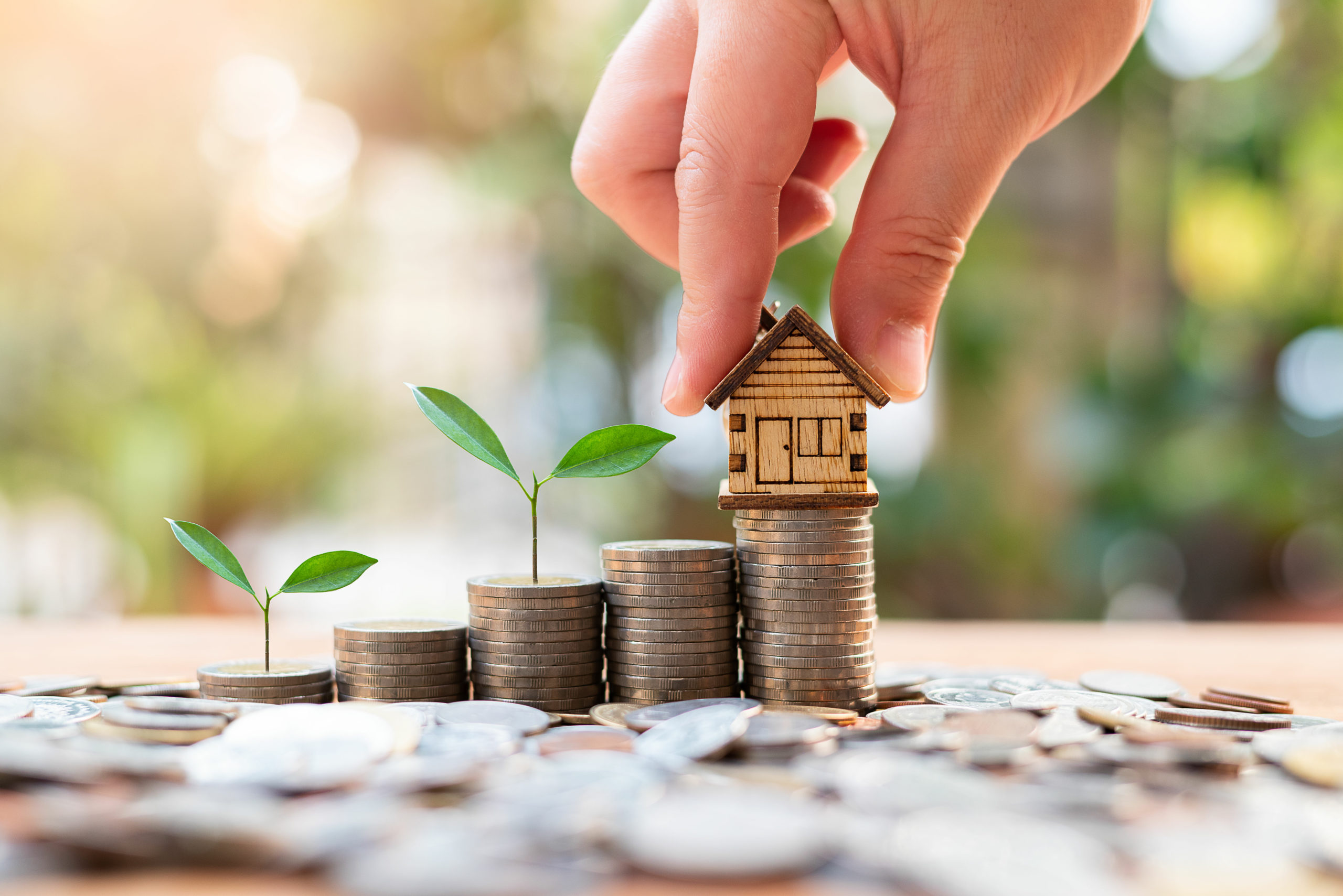 Hand protecting wooden house model on growing stacks of coins with green plants representing investment property growth and landlord insurance protection